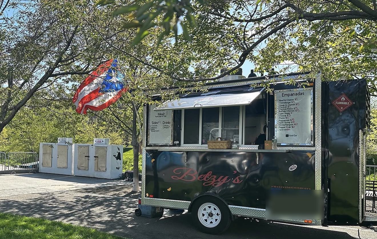 Betzy's food trailer parked under shady trees with a Puerto Rican flag flying